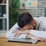 “늘 피곤한 당신, 만성피로 극복하는 ‘슬로우 라이프’ 바람” a man sitting at a desk with his head in his hands